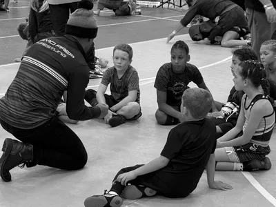 Coach kneeling and talking to a group of young children sitting on a gym floor during a wrestling event